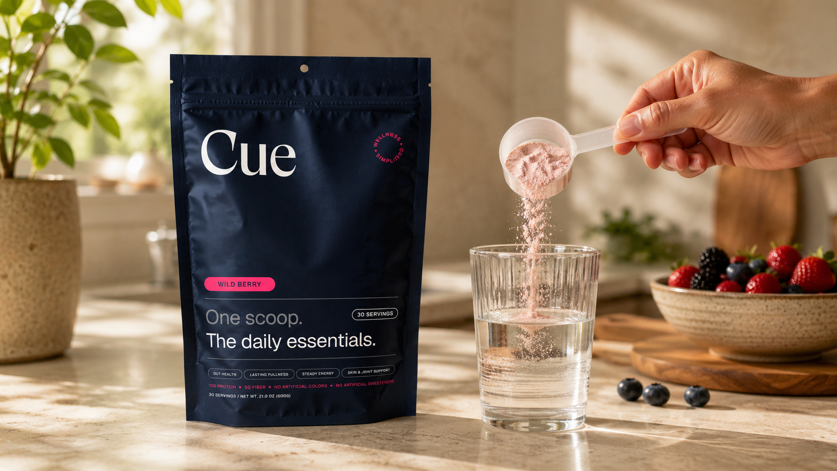 A scoop of Cue Wild Berry powder being poured into a glass of water beside the pouch